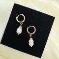 A pair of gold hoop earrings with dangling white baroque pearls, displayed on a black presentation box over a pale yellow satin background.