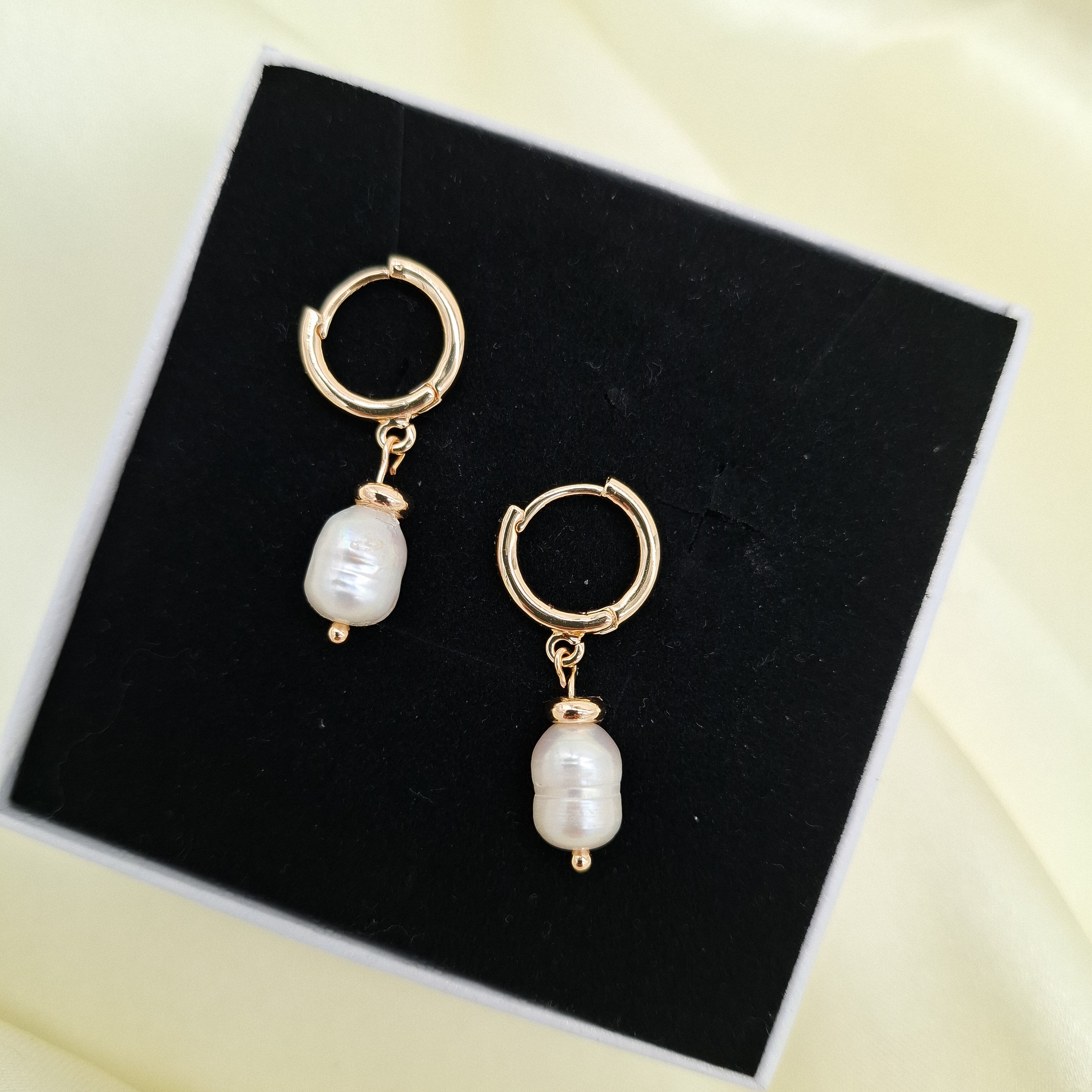 A pair of gold hoop earrings with dangling white baroque pearls, displayed on a black presentation box over a pale yellow satin background.