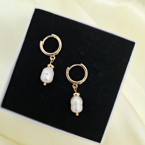 A pair of gold hoop earrings with dangling white baroque pearls, displayed on a black presentation box over a pale yellow satin background.