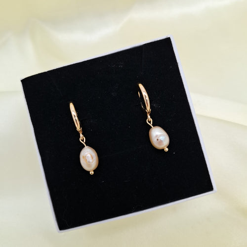 A pair of gold drop earrings with irregular white pearls, displayed on a black square jewelry box against a soft cream satin background.
