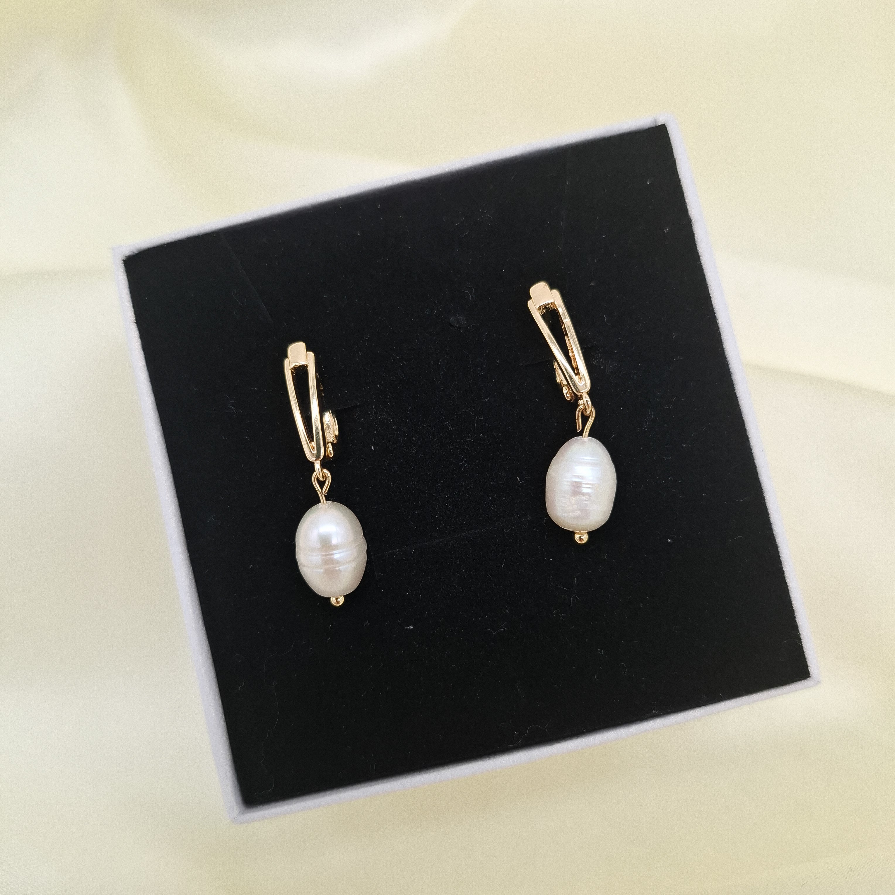freshwater pearl drop earrings with gold-tone huggie hoop clasps, displayed on a black velvet jewelry box against a cream fabric background