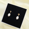 A pair of gold drop earrings with irregular white freshwater pearls, displayed on a black jewelry box against a soft cream-colored fabric background.