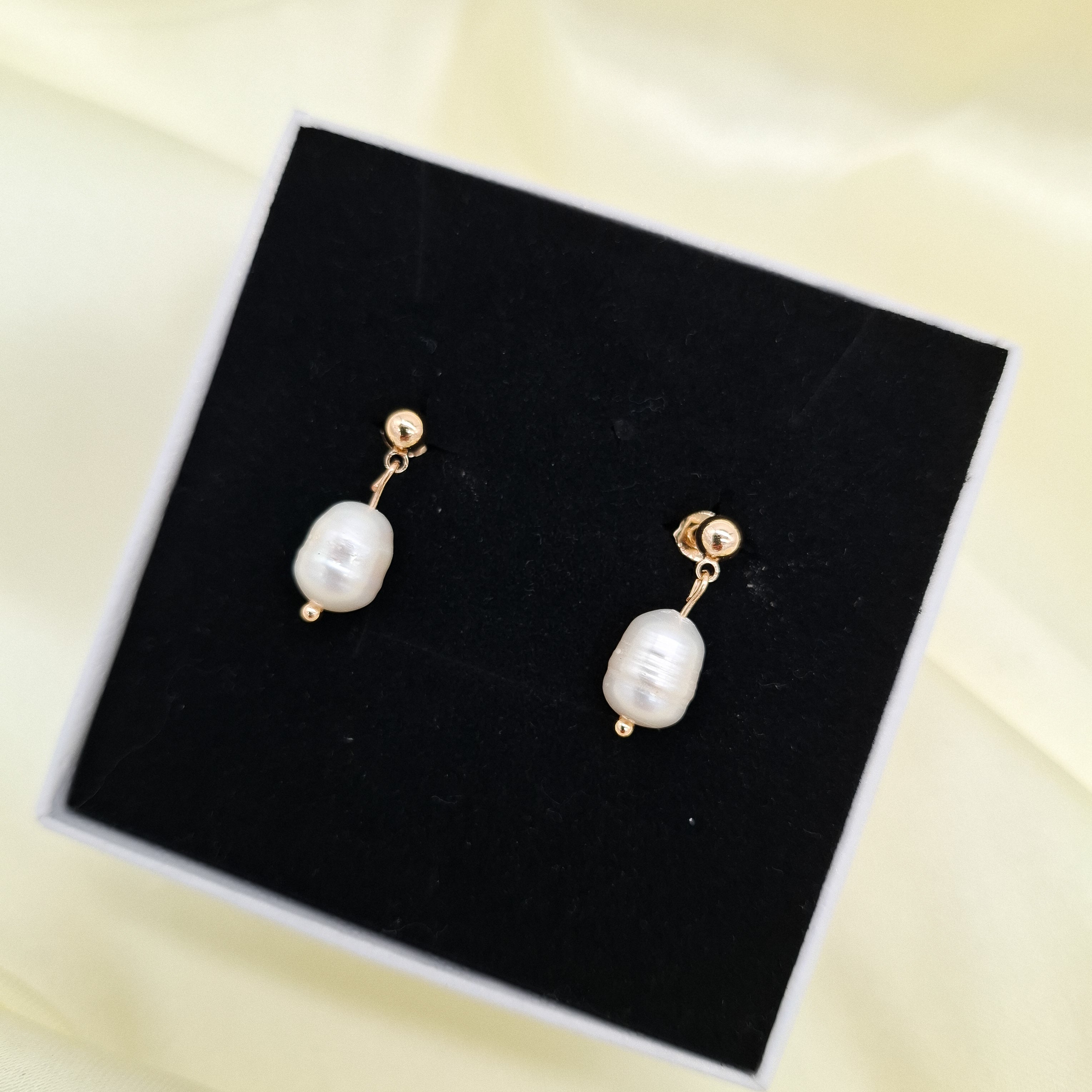 A pair of gold drop earrings with irregular white freshwater pearls, displayed on a black jewelry box against a soft cream-colored fabric background.